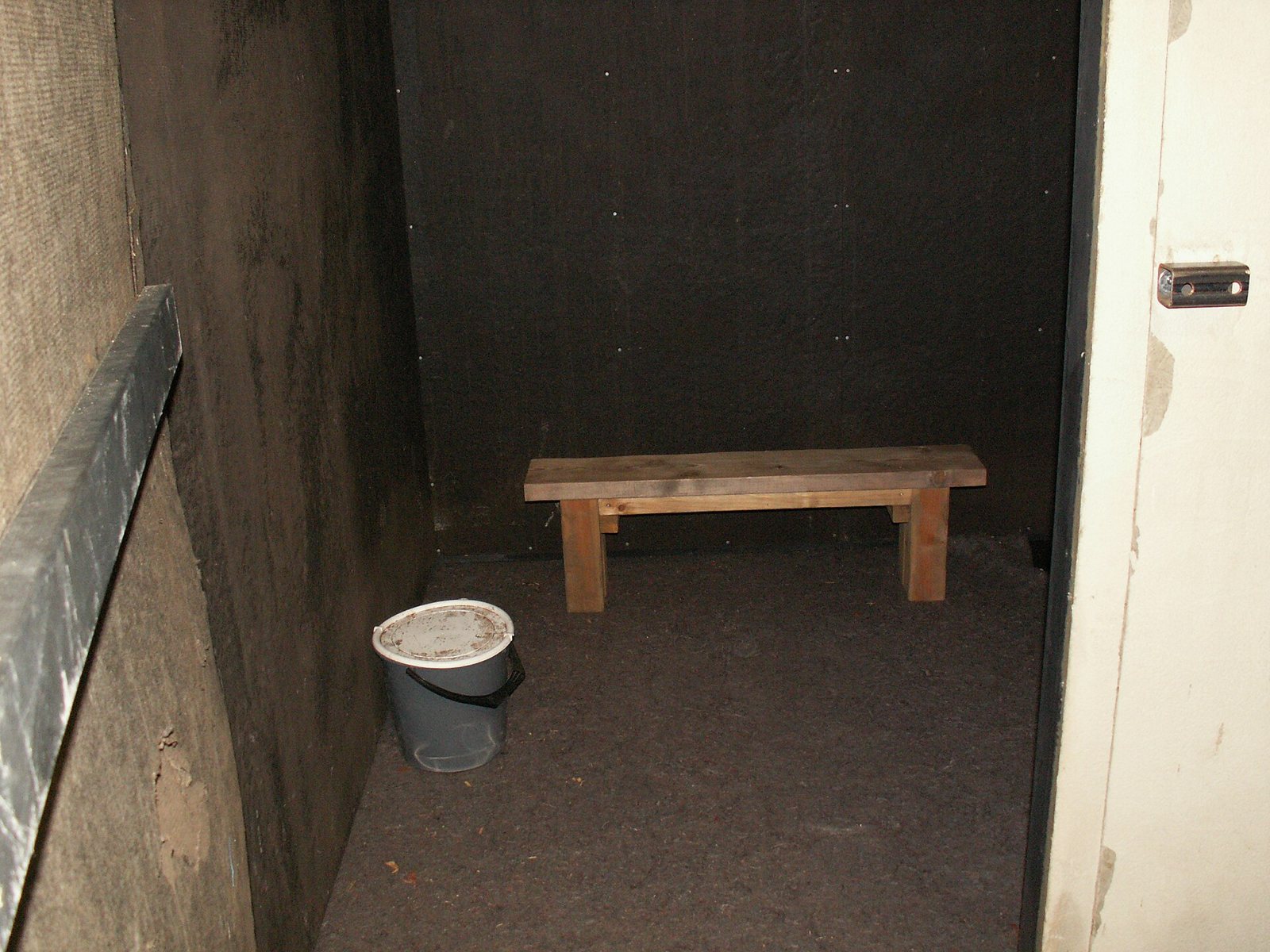 The inside of a plain wooden replica cell with a bench and a bucket